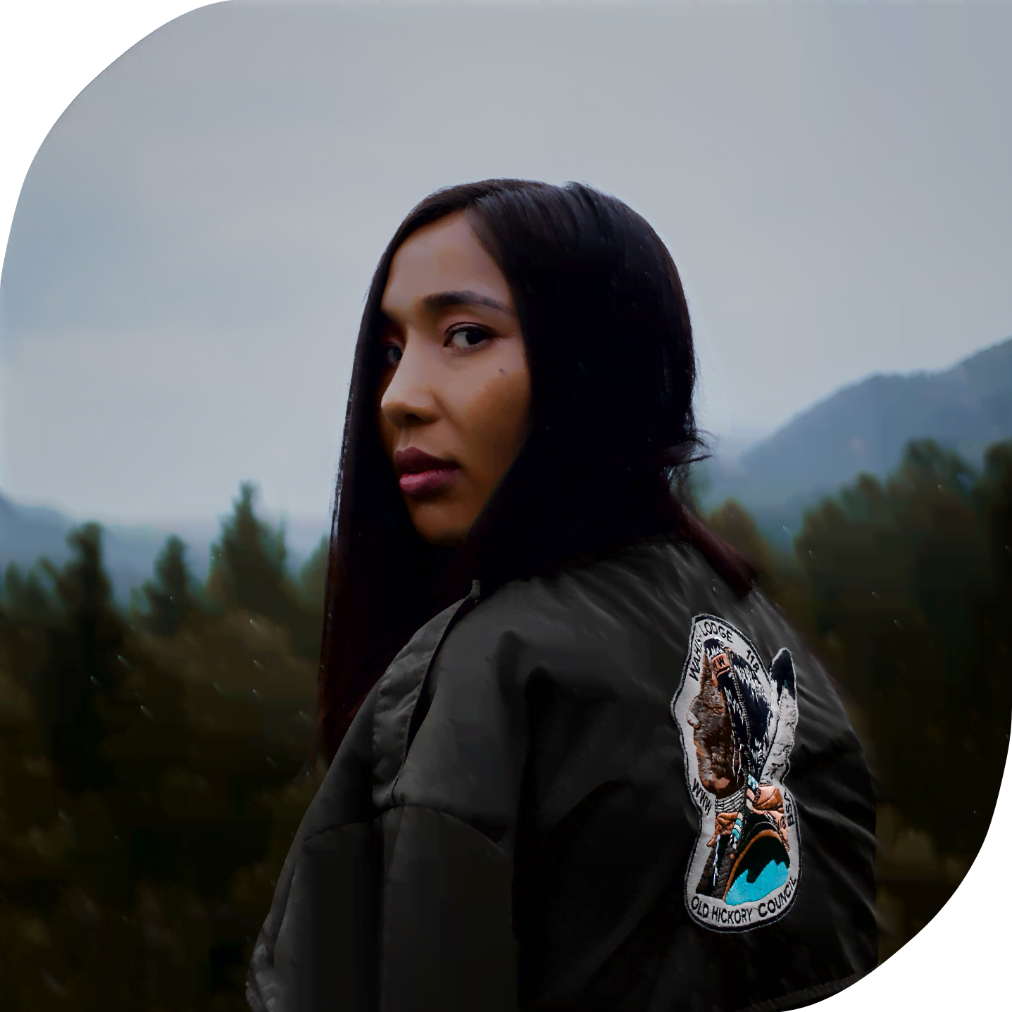 A young Indigenous woman glances over her shoulder on a cloudy day. Behind her are mountains and a forest.