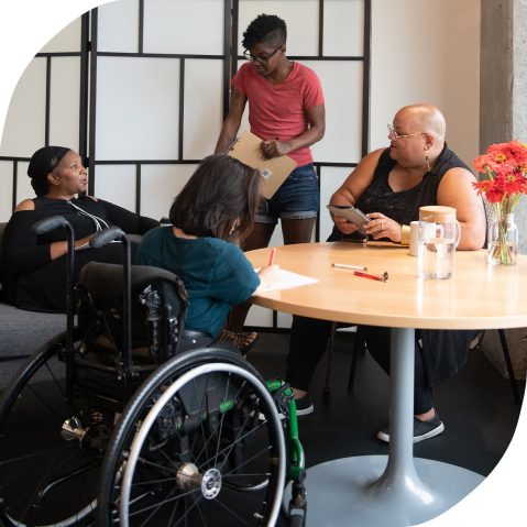 Four disabled people of color gather around a table during a meeting. Photo by affecttheverb.com/disabledandhere