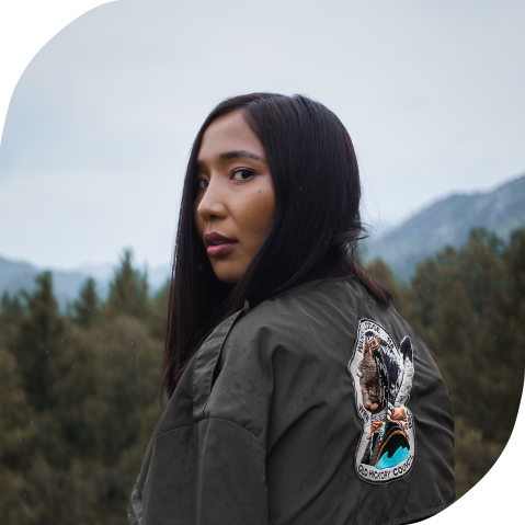Inquiry document banner A young Indigenous woman glances over her shoulder on a cloudy day. Behind her are mountains and a forest.
